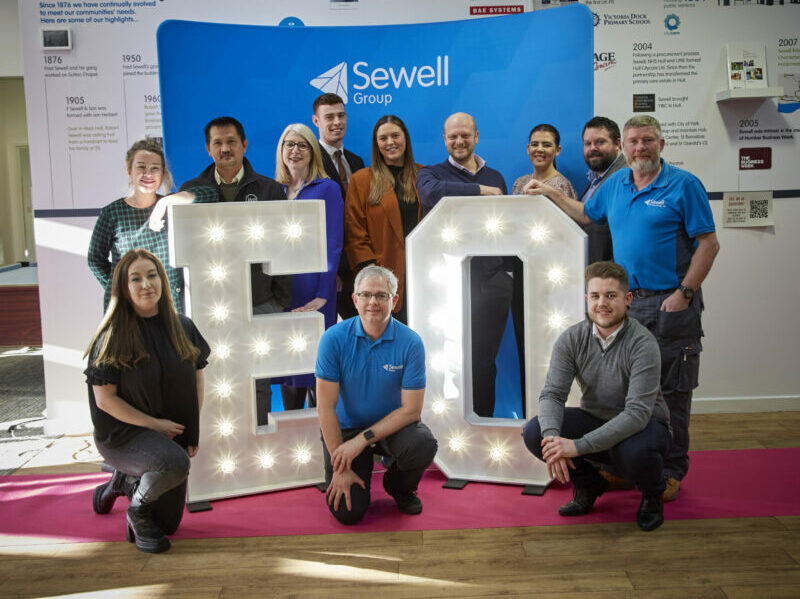 A group of Sewell Group staff stand around two large lit-up letters E and O