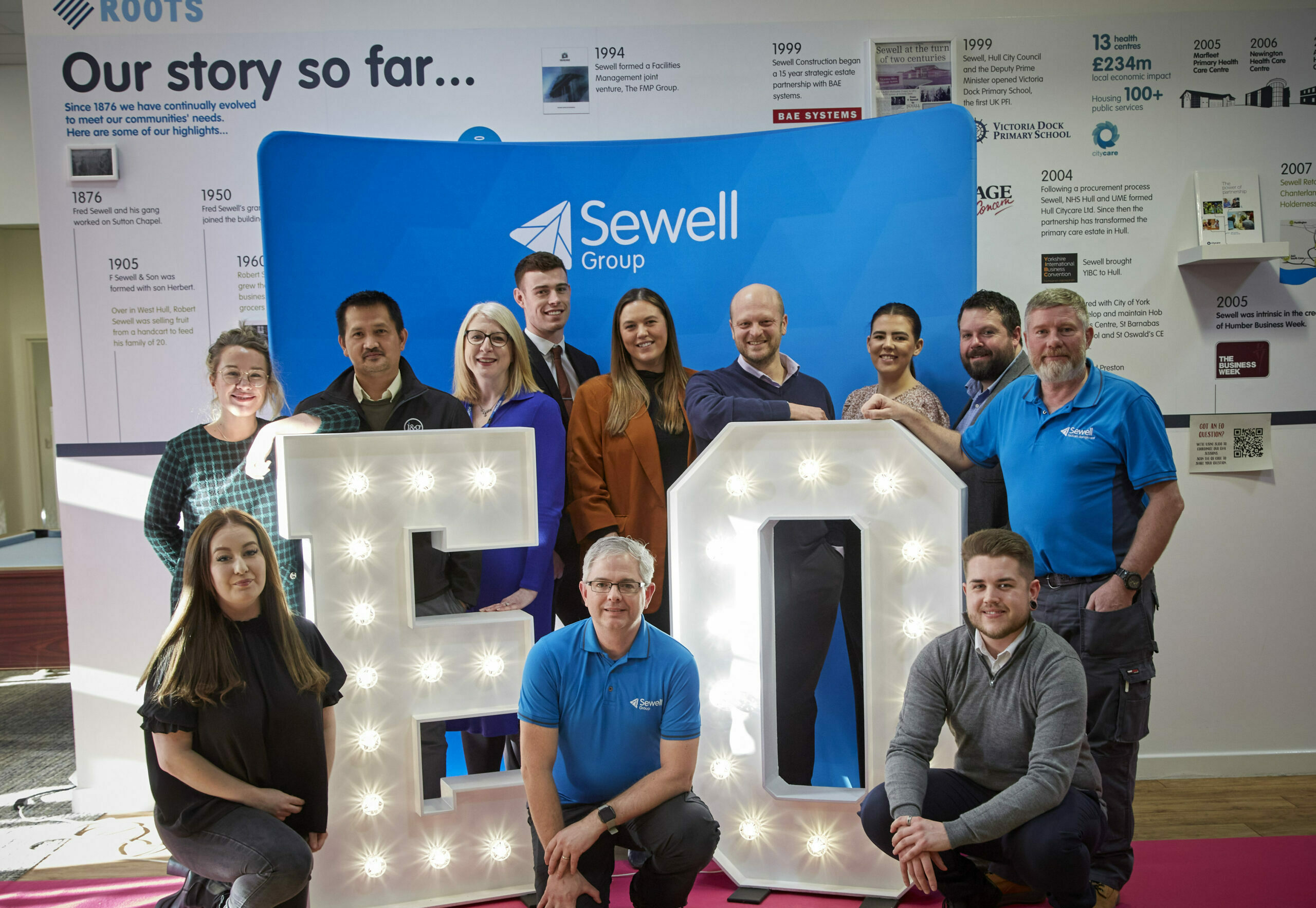 A group of Sewell Group staff stand around two large letters, E and O